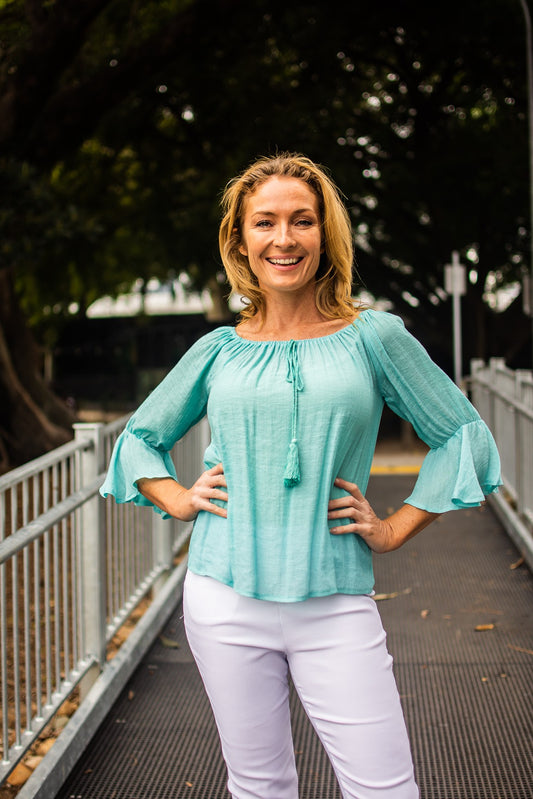 Woman standing on a pedestrian bridge in a power pose with hands on hips. She's wearing an aqua round-neck Bell Top with decorative tassel drawstrings and 3/4 fill sleeves, and white pants. There are trees in the background.