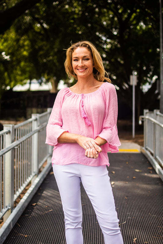 Woman in a pink Bell Top with 3/4 sleeves and tassel drawstring round neckline, and white pants standing on a pedestrian pathway with trees in the background