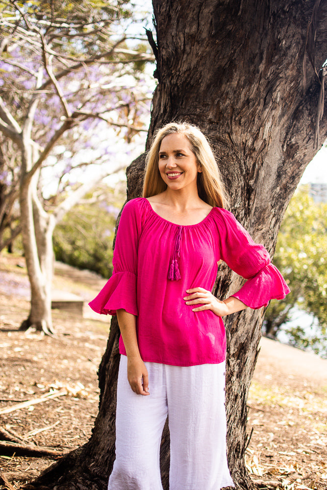 Woman in a hot pink Bell Top with 3/4 sleeves and tassel drawstring round neckline, and white beach pants near a tree with blooming jacaranda trees in the background