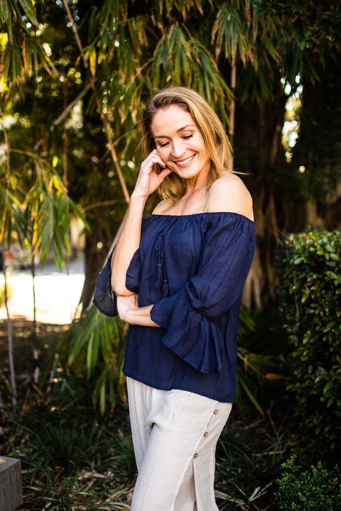 Woman in a navy Bell Top with 3/4 sleeves and tassel drawstring round neckline. She is wearing the bell top in the off the shoulder style, and with beige beach pants. Gardens and trees in the background
