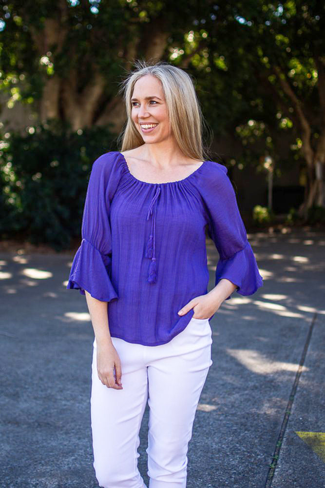 Woman in a purple Bell Top with 3/4 sleeves and tassel drawstring round neckline, and white pants standing on a driveway with gardens and trees in the background