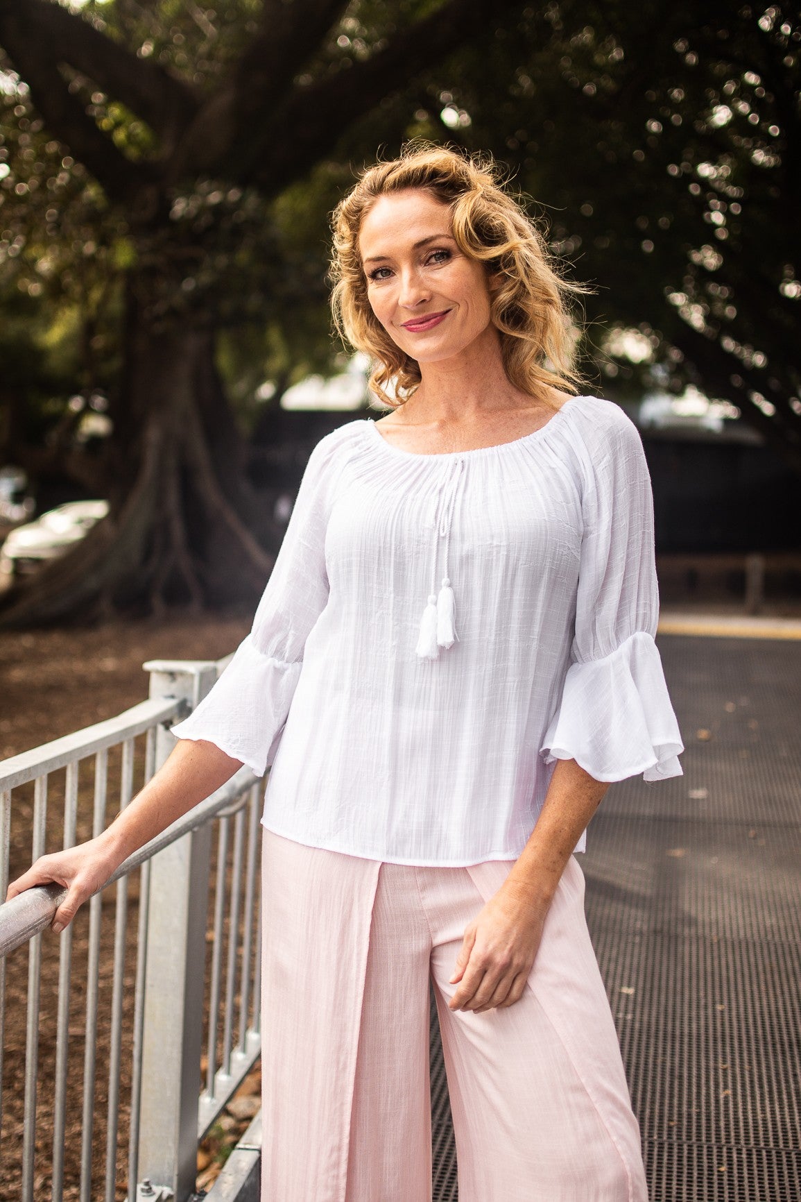 Woman in a white Bell Top with 3/4 sleeves and tassel drawstring round neckline, and baby pink pants standing on a pedestrian pathway with trees in the background