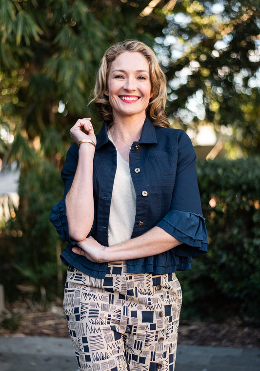 Woman wearing a navy Frill Denim Jacket over a beige top and beige and navy patterned pants with a bushes and trees background