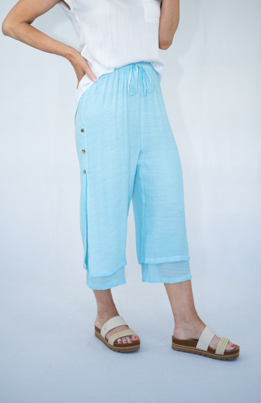 Woman wearing baby blue Beach Pants with a white top and beach sandals. Studio white background.