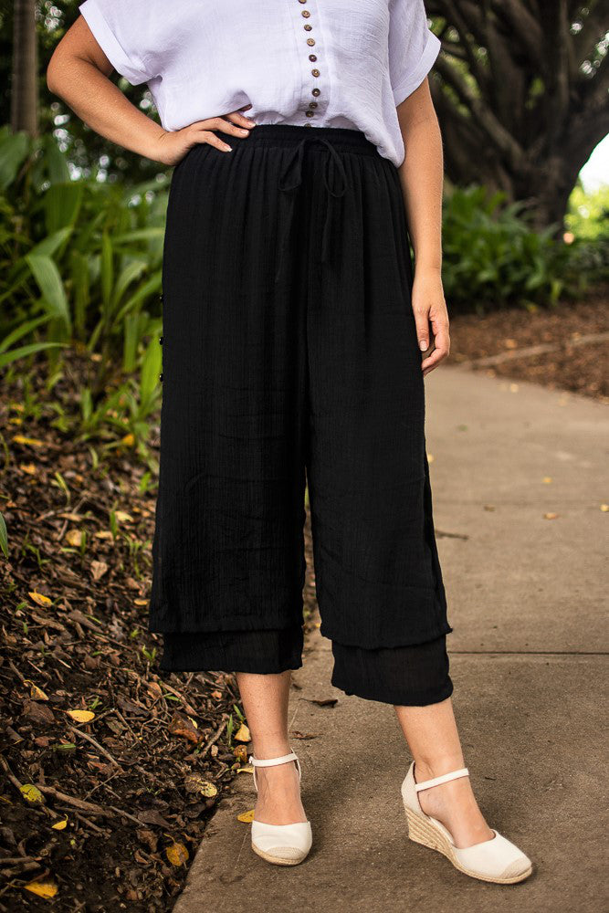 A person wearing black Beach Pants with an elastic waistband and wooden side buttons, paired with a white top and white wedge shoes, standing on a pavement with tended gardens and trees in the background.