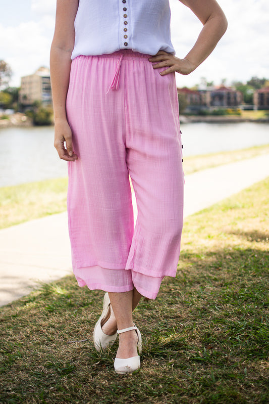 A person wearing pink Beach Pants with an elastic waistband and wooden side buttons, paired with a white top and white shoes, standing on grass with a body of water and buildings in the background.