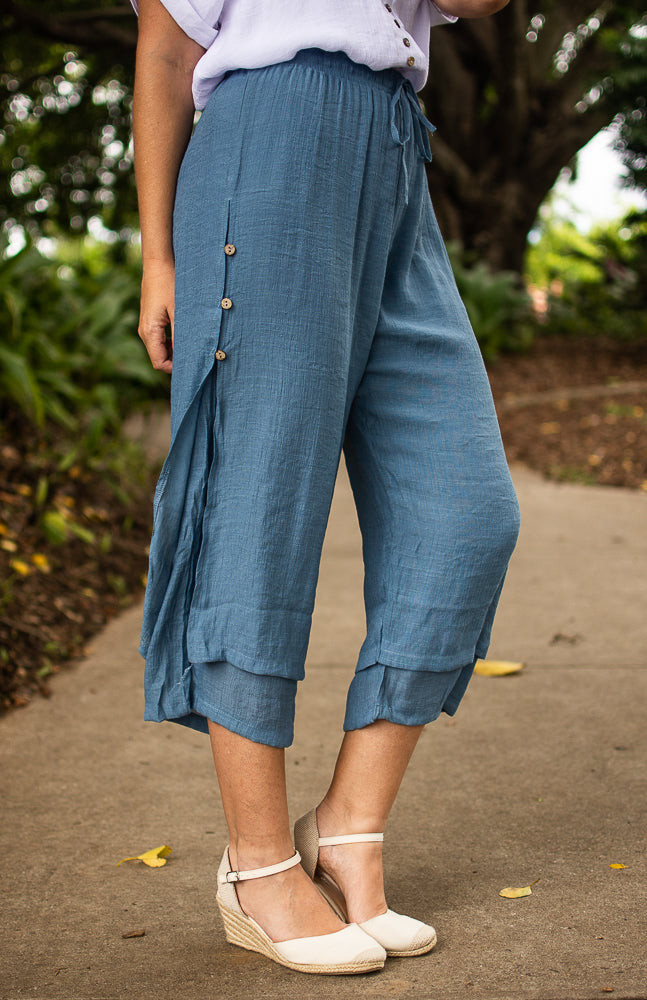 A person wearing denim Beach Pants with an elastic waistband and wooden side buttons, paired with a white top and white wedge shoes, standing on a pavement with tended gardens and trees in the background.