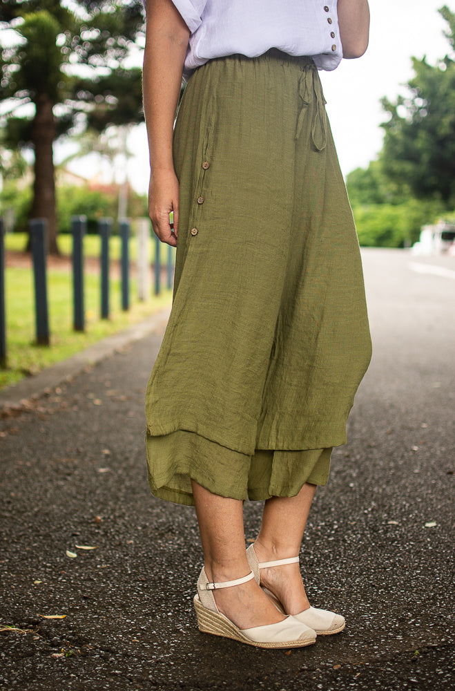 A person wearing khaki Beach Pants with an elastic waistband and wooden side buttons, paired with a white top and white wedge shoes, standing on a pavement with tended gardens and trees in the background.