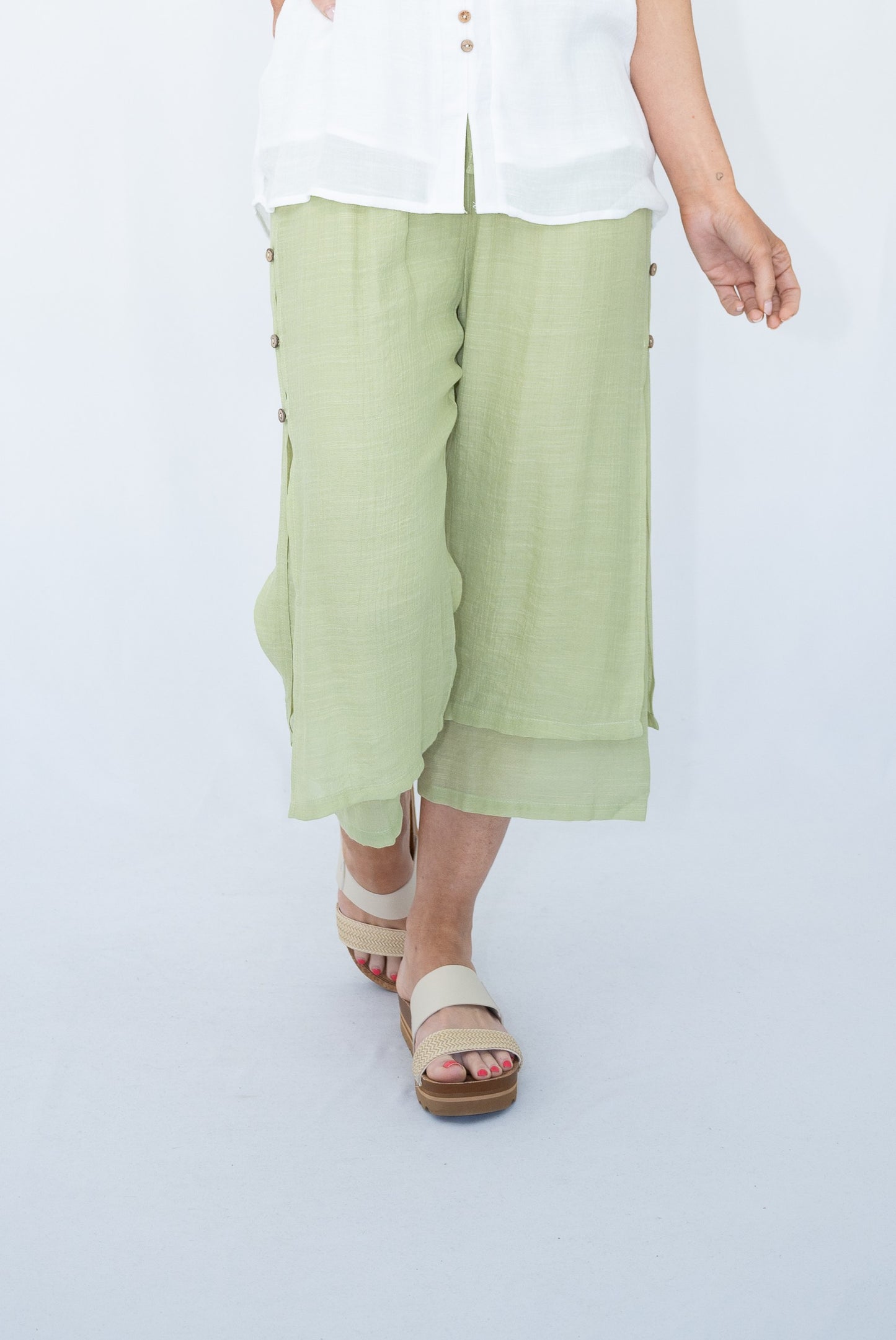Woman wearing light sage Beach Pants with a white top and beach sandals. Studio white background.