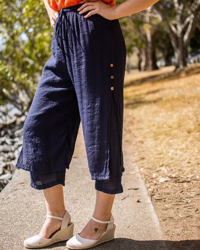 A person wearing navy Beach Pants with an elastic waistband and wooden side buttons, paired with an orange top and white wedge shoes, standing on a pavement with trees and the riverside in the background.