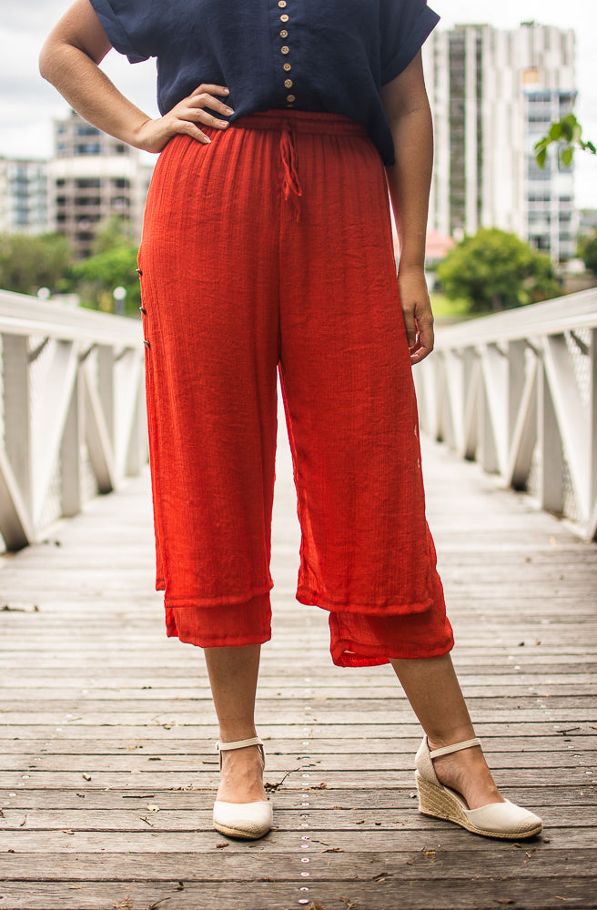 A person wearing rust Beach Pants with an elastic waistband and wooden side buttons, paired with a navy top and white wedge shoes, standing on a jetty with trees and city in the background.