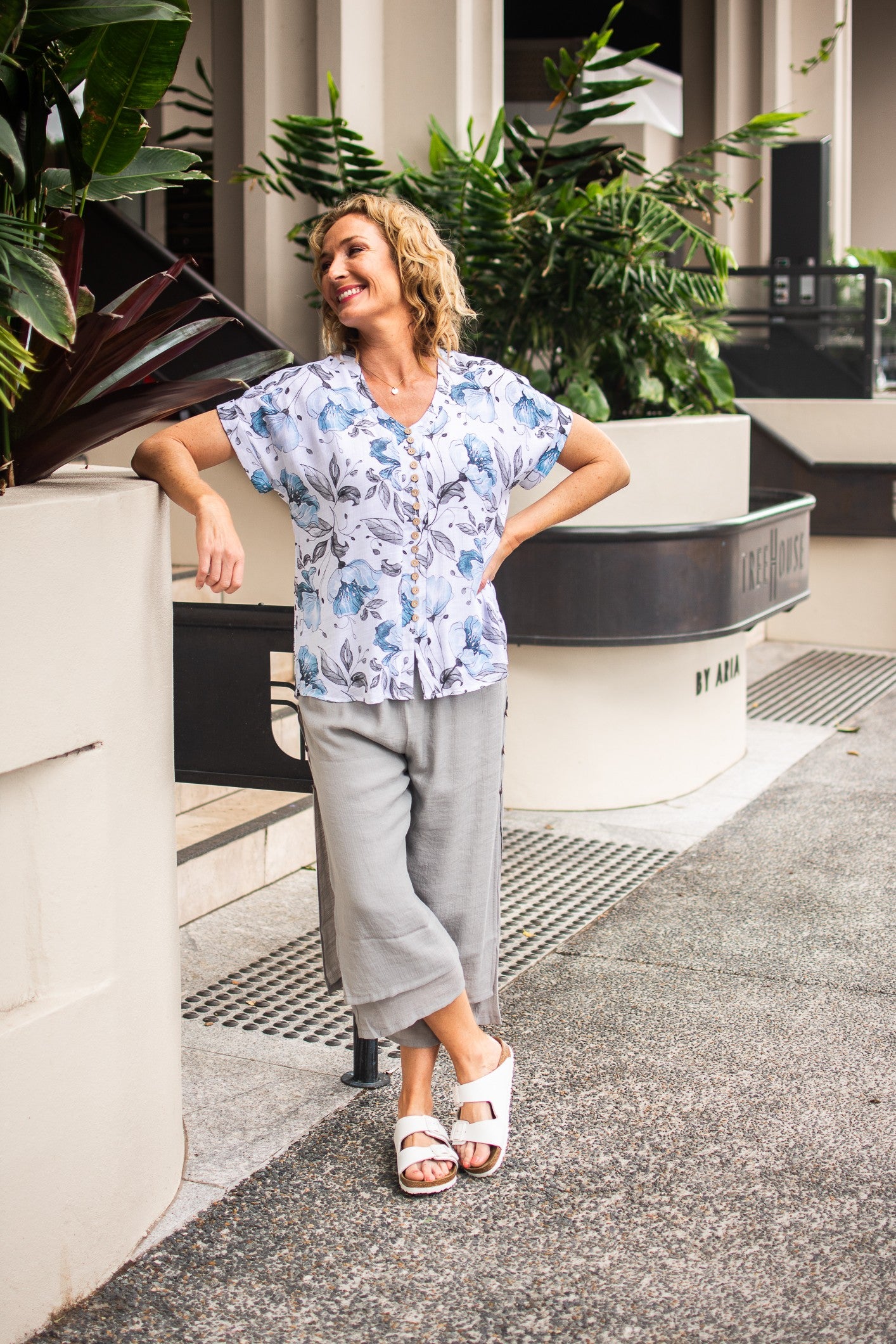 A woman wearing silver Beach Pants with an elastic waistband and wooden side buttons, paired with a floral top and white shoes, standing outside a building on pebble pavement with stairs and gardens in the background.