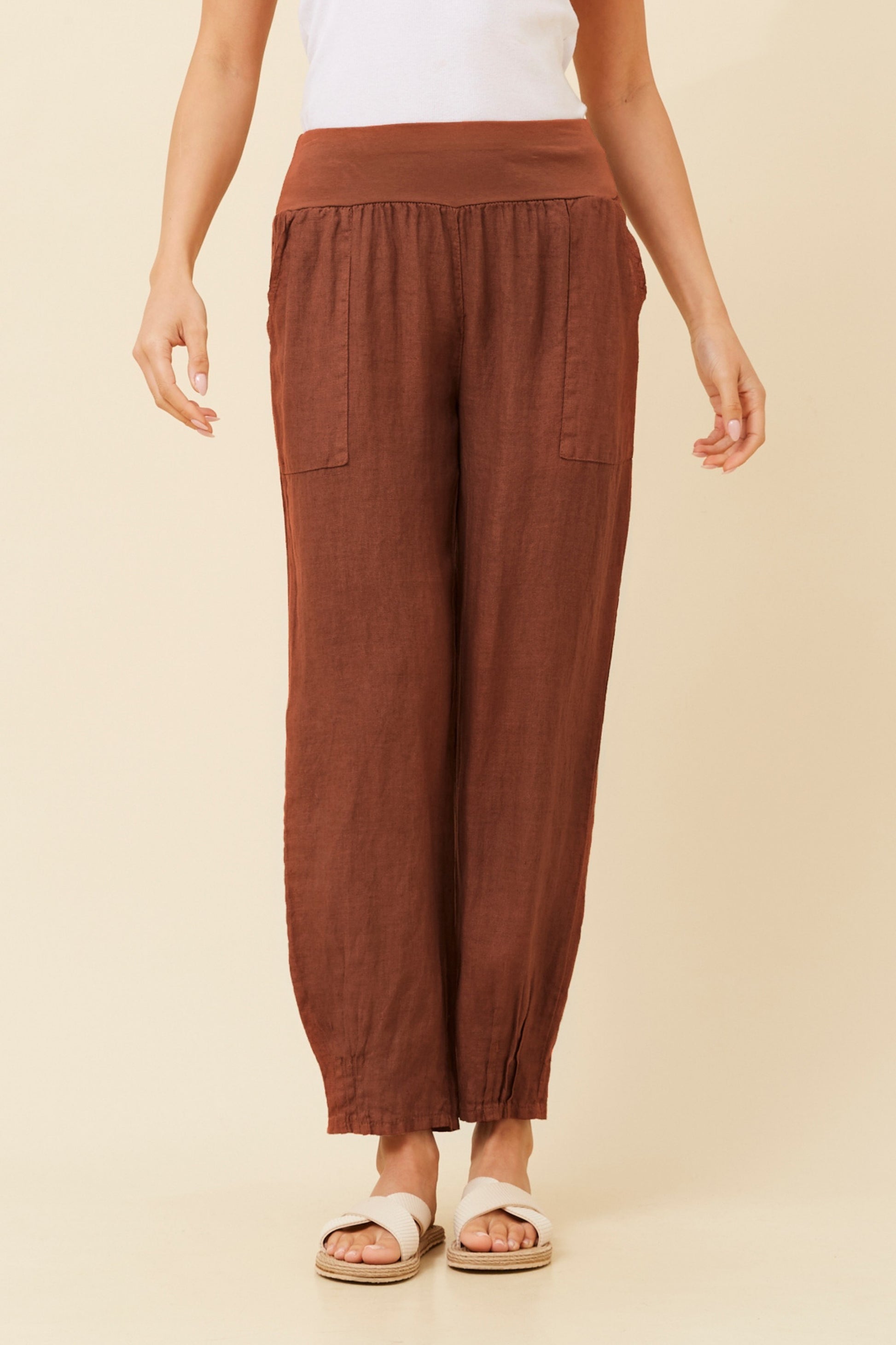 Woman wearing rust coloured Linen Italian Pants with an adjustable waistband, front pockets, and pleated hems, paired with a white top and white criss-cross shoes. Cream studio background.
