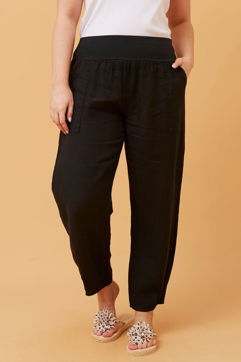 Woman wearing black Linen Italian Pants with an adjustable waistband, front pockets, and pleated hems, paired with a white top and fluffy patterned shoes. Orange studio background.