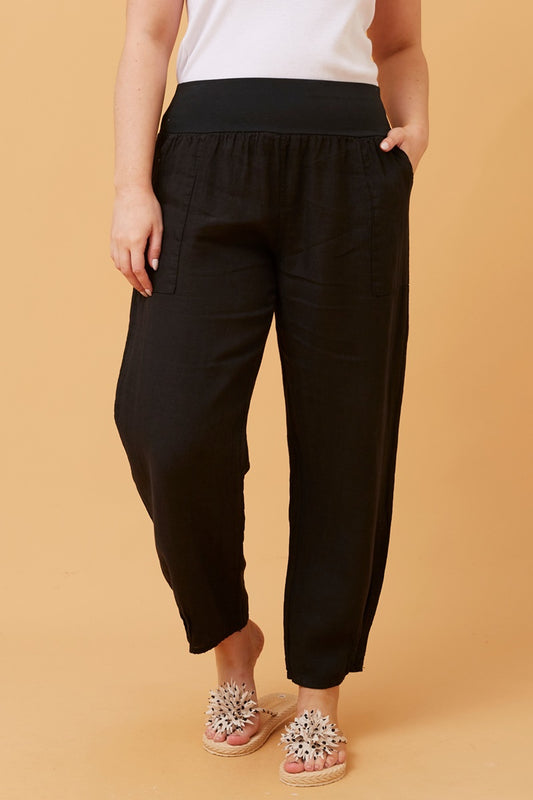Woman wearing black Linen Italian Pants with an adjustable waistband, front pockets, and pleated hems, paired with a white top and fluffy patterned shoes. Orange studio background.