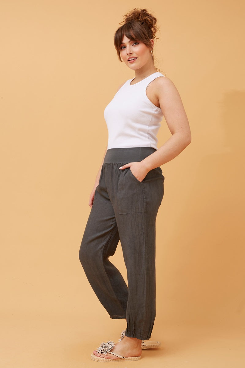 Side view of woman wearing charcoal Linen Italian Pants with an adjustable waistband, front pockets, and pleated hems, paired with a white top and fluffy patterned shoes. Orange studio background.