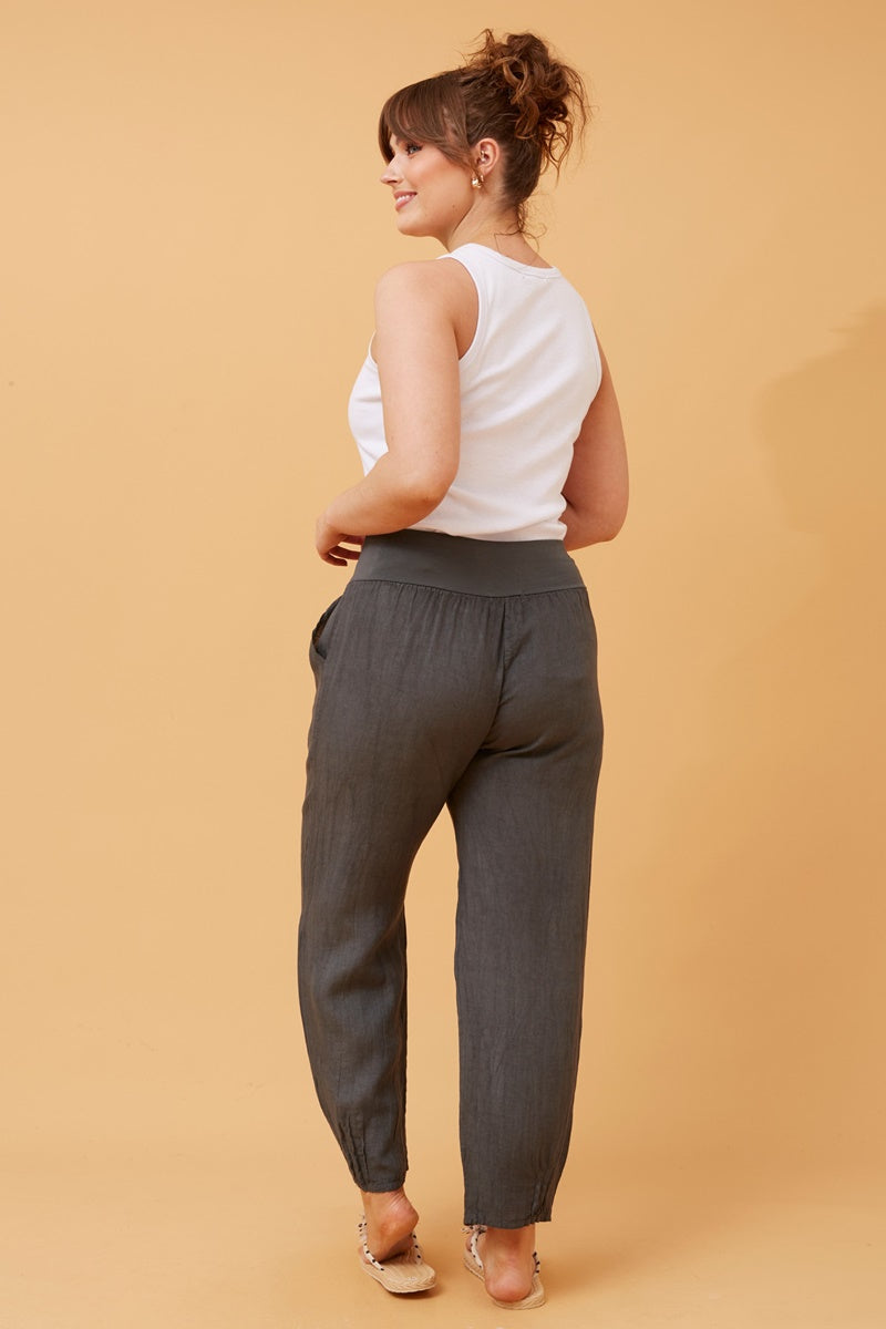 Back view of woman wearing charcoal Linen Italian Pants with an adjustable waistband, front pockets, and pleated hems, paired with a white top and fluffy patterned shoes. Orange studio background.