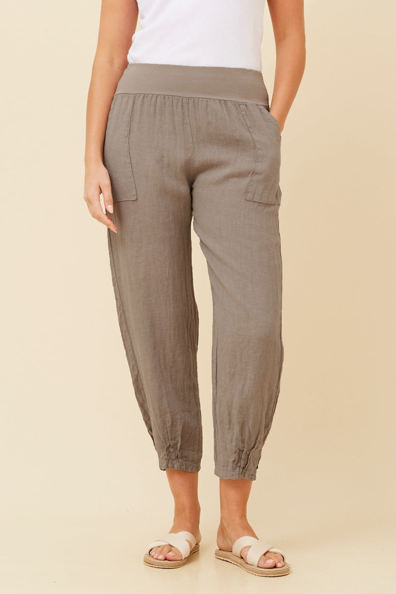Woman wearing fango coloured Linen Italian Pants with an adjustable waistband, front pockets, and pleated hems, paired with a white top and white criss-cross shoes. Cream studio background.