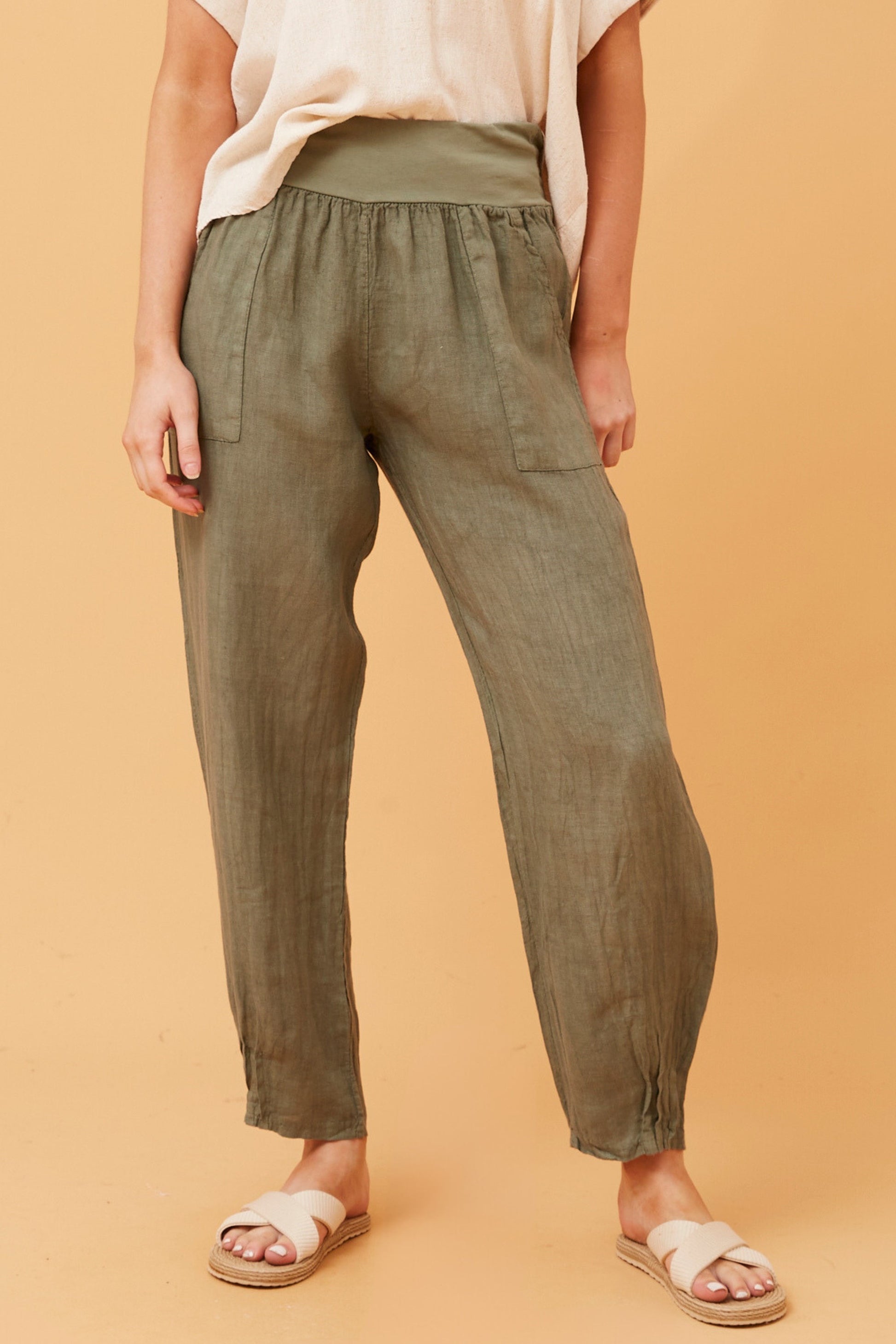 Woman wearing khaki Linen Italian Pants with an adjustable waistband, front pockets, and pleated hems, paired with a beige top and white criss-cross shoes. Orange studio background.