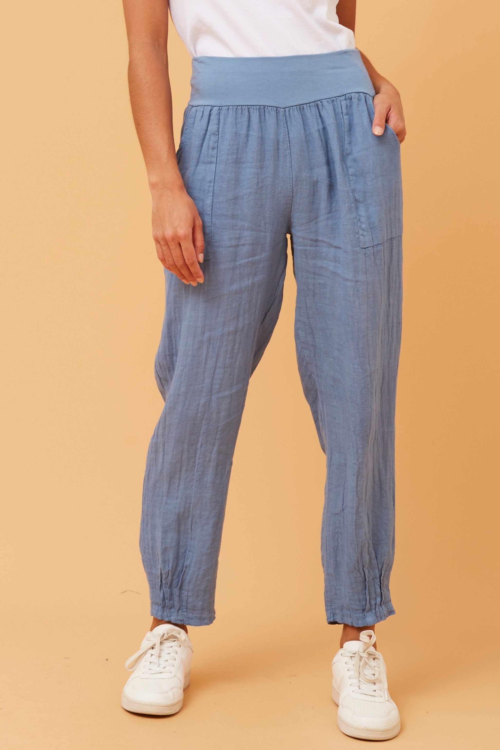 Woman wearing denim Linen Italian Pants with an adjustable waistband, front pockets, and pleated hems, paired with a white top and white runner shoes. Orange studio background.