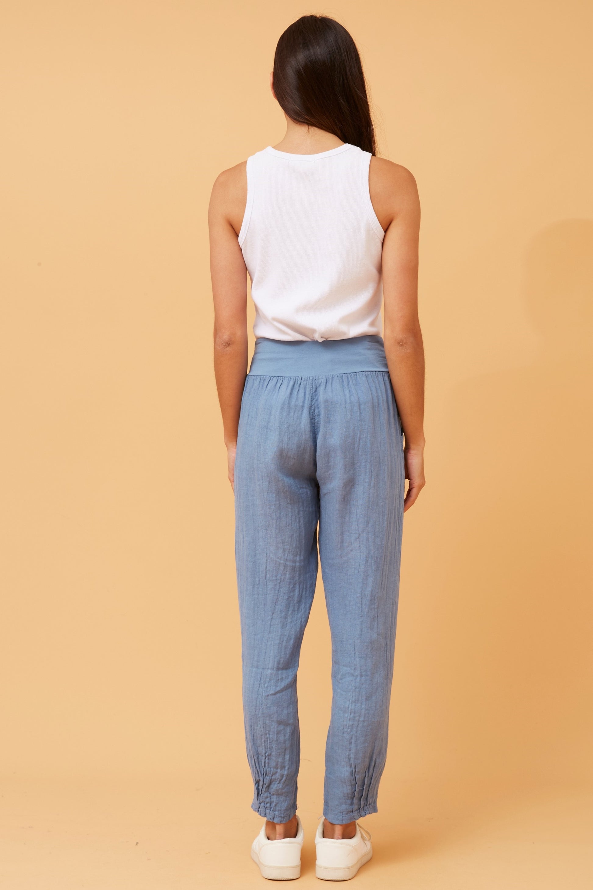 Back view of woman wearing denim Linen Italian Pants with an adjustable waistband, front pockets, and pleated hems, paired with a white top and white runner shoes. Orange studio background.