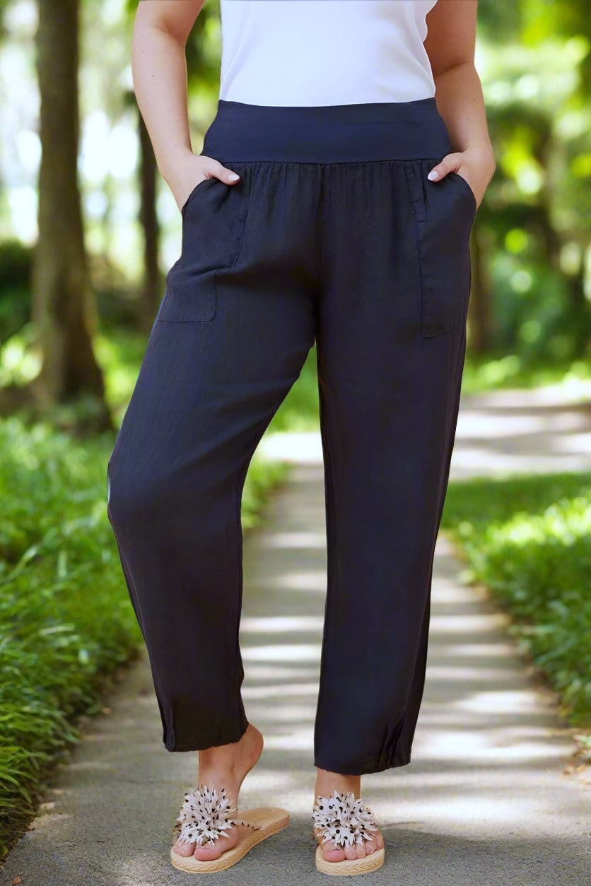 Woman wearing navy Linen Italian Pants with an adjustable waistband, front pockets, and pleated hems, paired with a white top and fluffy patterned shoes. AI generated pathway through city park background.