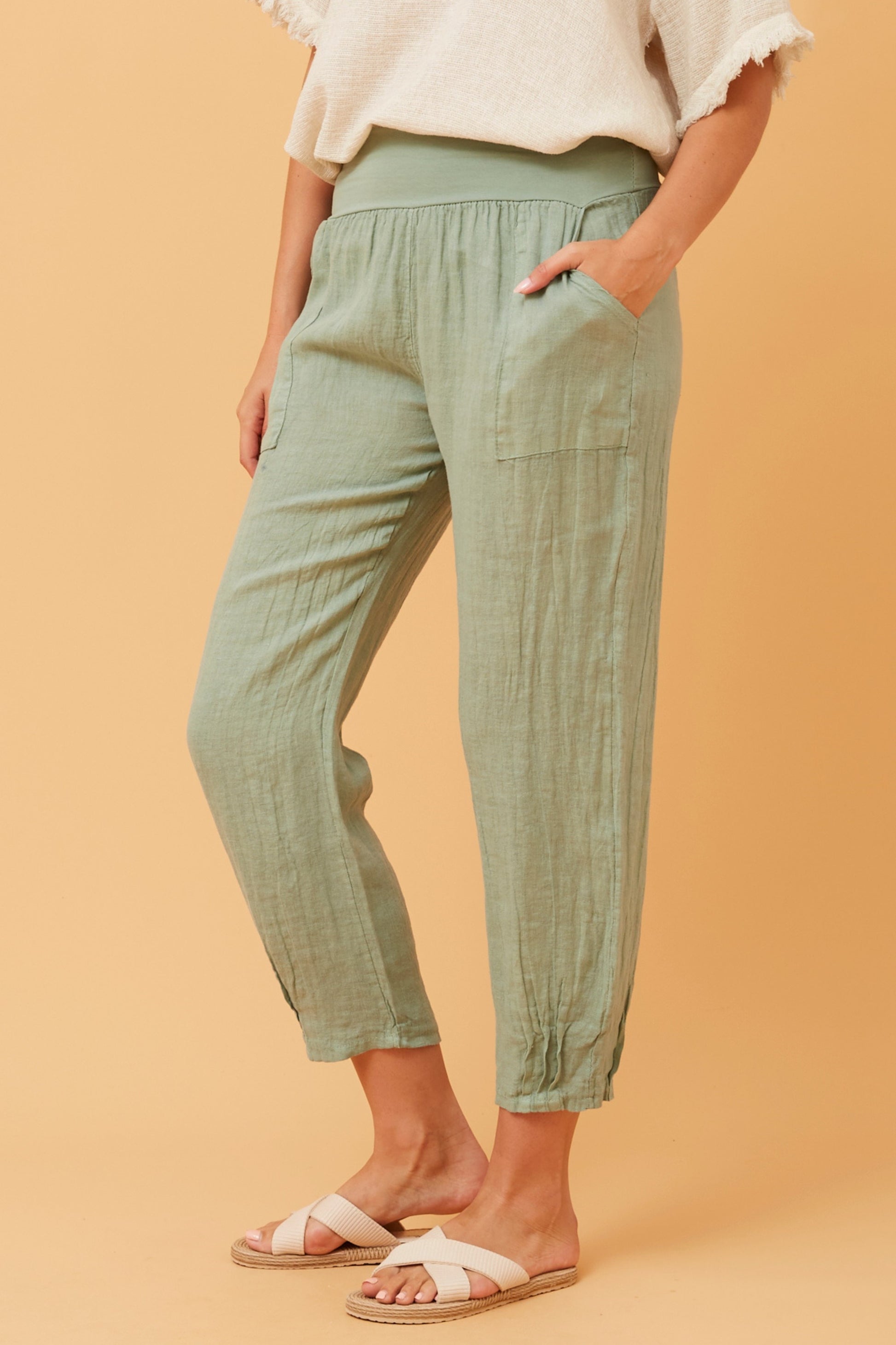 Woman wearing sage Linen Italian Pants with an adjustable waistband, front pockets, and pleated hems, paired with a beige top and beige criss-cross shoes. Orange studio background.
