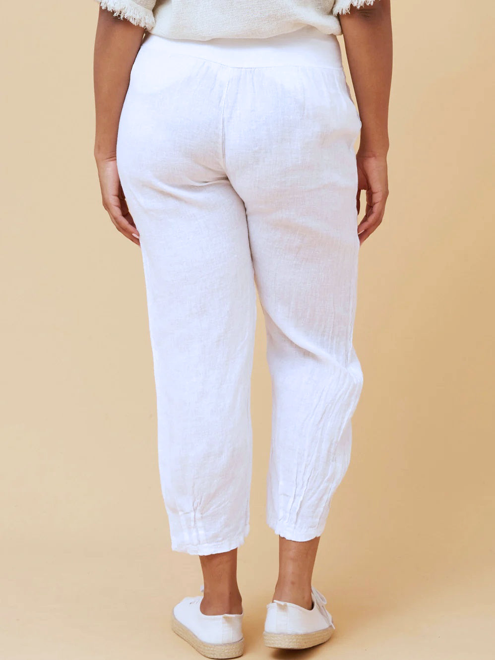 Woman wearing white Linen Italian Pants with an adjustable waistband, front pockets, and pleated hems, paired with a beige top and white runner shoes. Orange studio background.