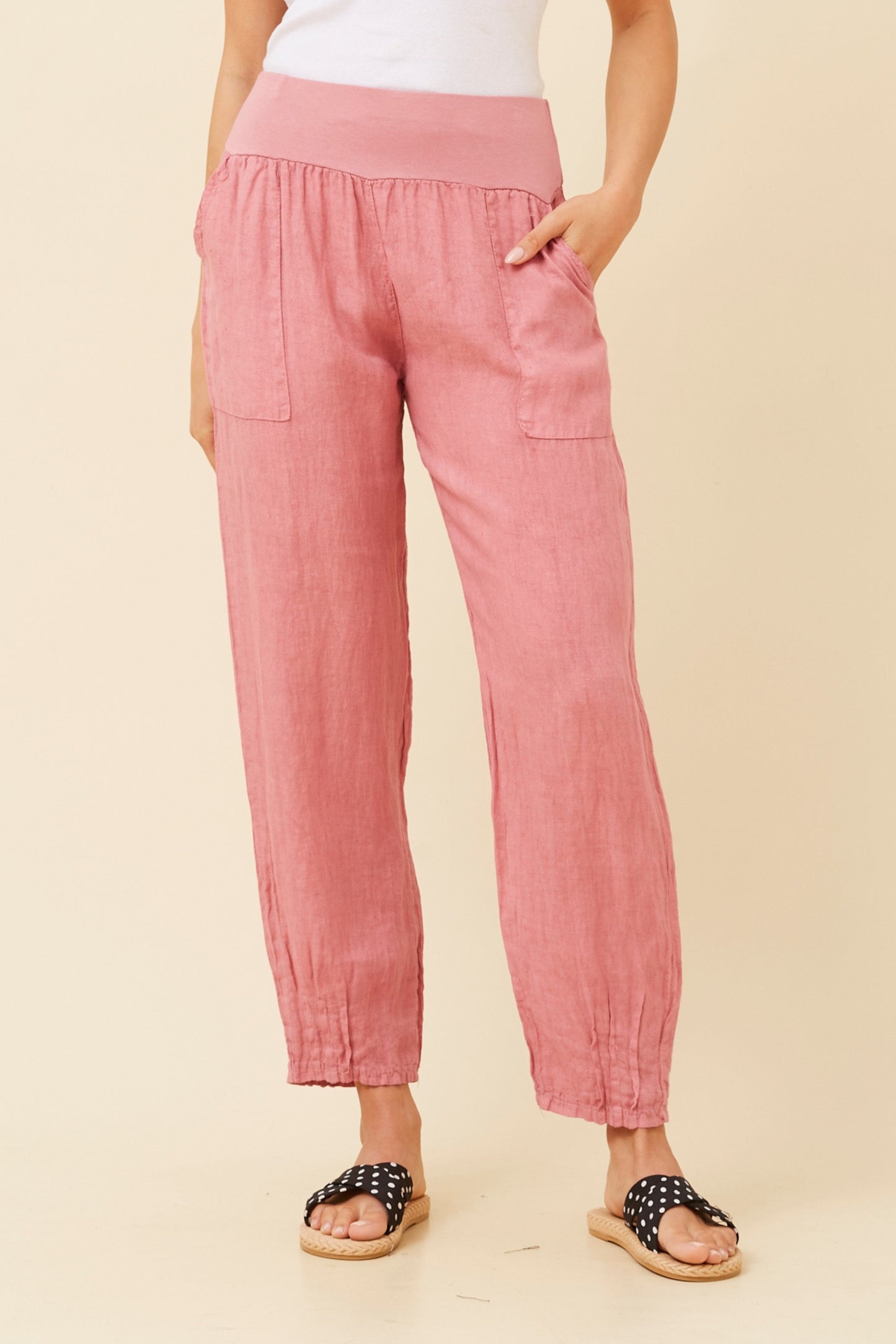 Woman wearing cipolla (or pink) Linen Italian Pants with an adjustable waistband, front pockets, and pleated hems, paired with a white top and pola dot patterned shoes. Beige studio background.