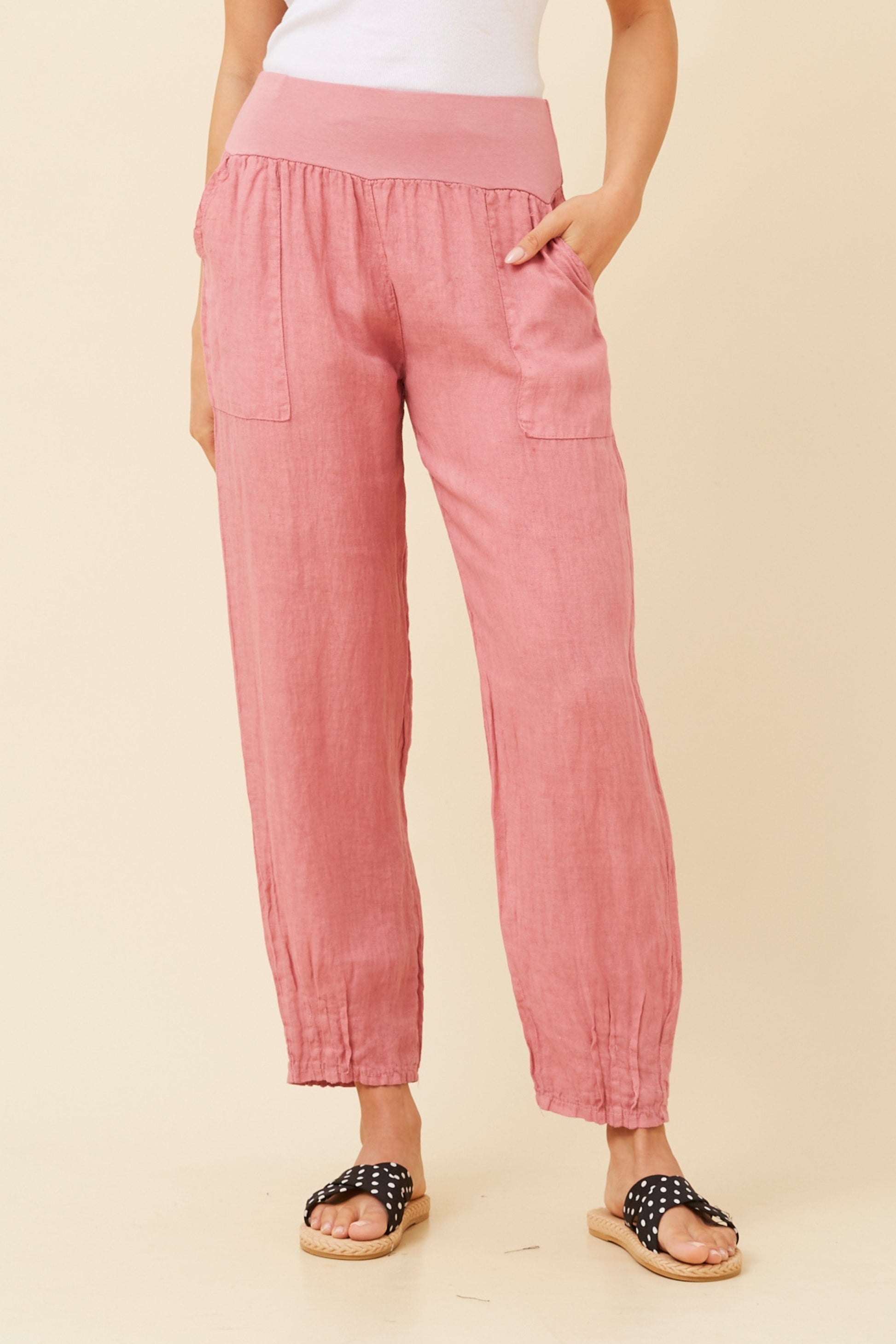 Woman wearing cipolla (or pink) Linen Italian Pants with an adjustable waistband, front pockets, and pleated hems, paired with a white top and pola dot patterned shoes. Beige studio background.