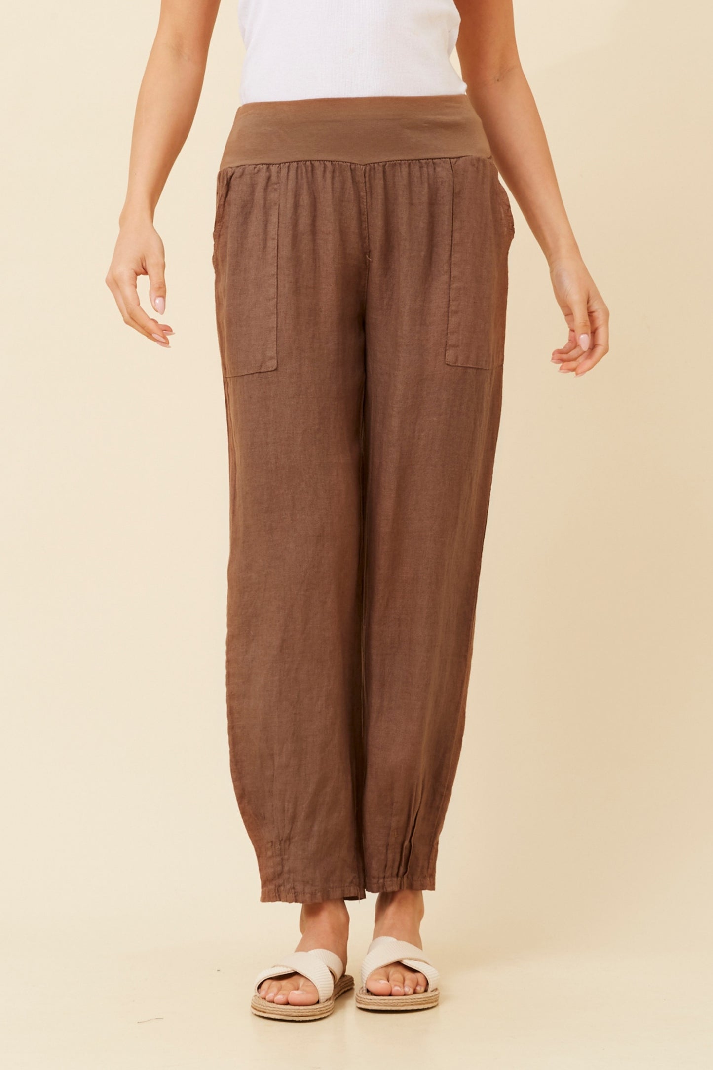 Woman wearing tobacco (or brown) Linen Italian Pants with an adjustable waistband, front pockets, and pleated hems, paired with a white top and white criss-cross shoes. Beige studio background.