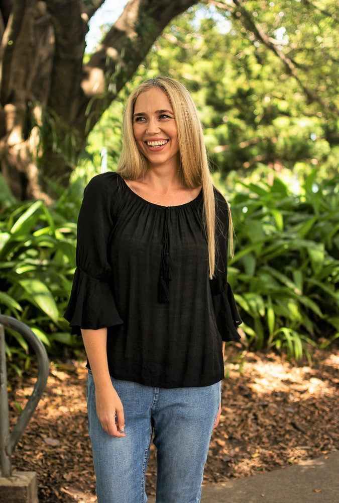 Woman in a black Bell Top with 3/4 sleeves and tassel drawstring round neckline, and blue jeans standing on a pathway with gardens and trees in the background