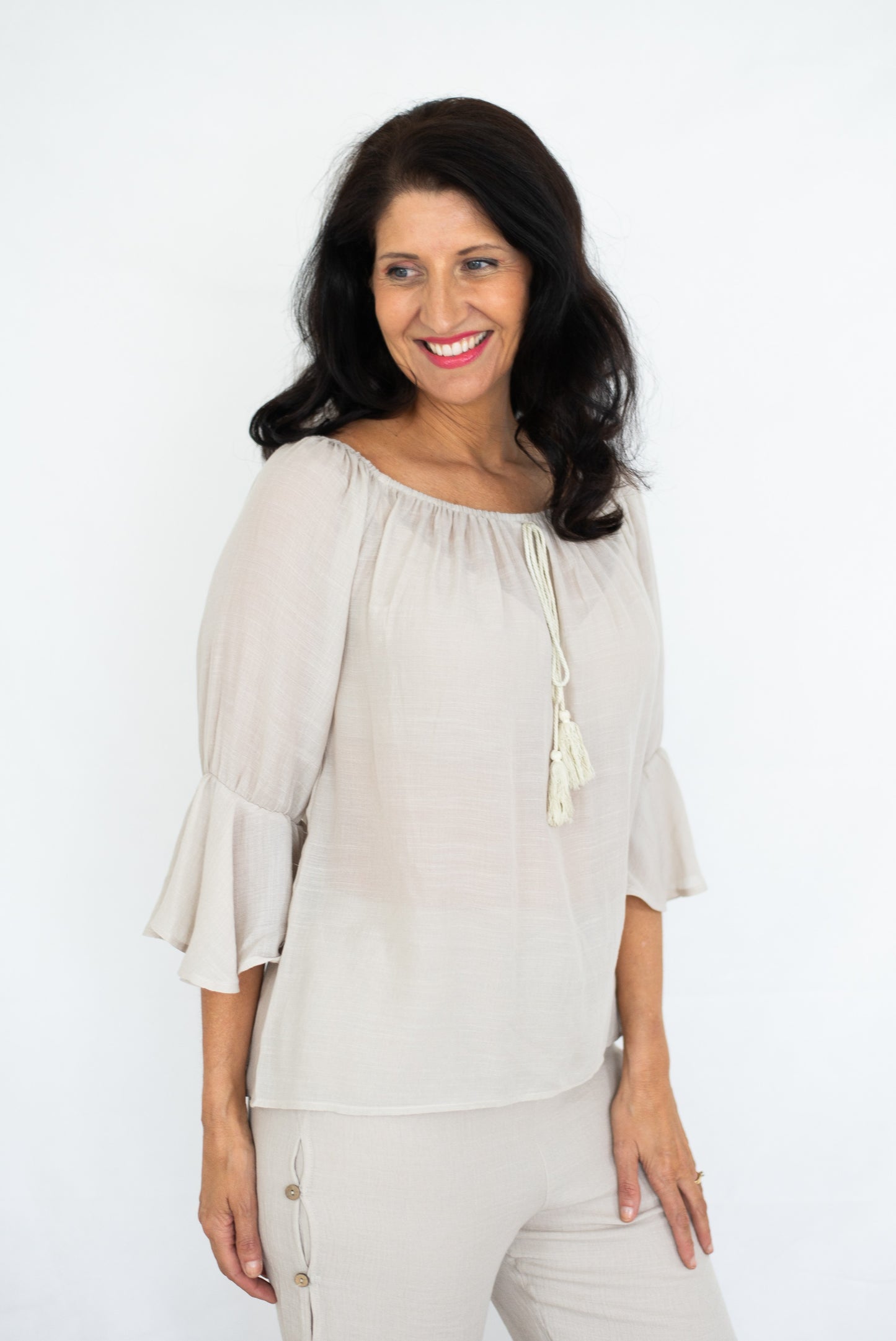Woman in a beige Bell Top with 3/4 sleeves and tassel drawstring round neckline, and beige beach pants against a white background