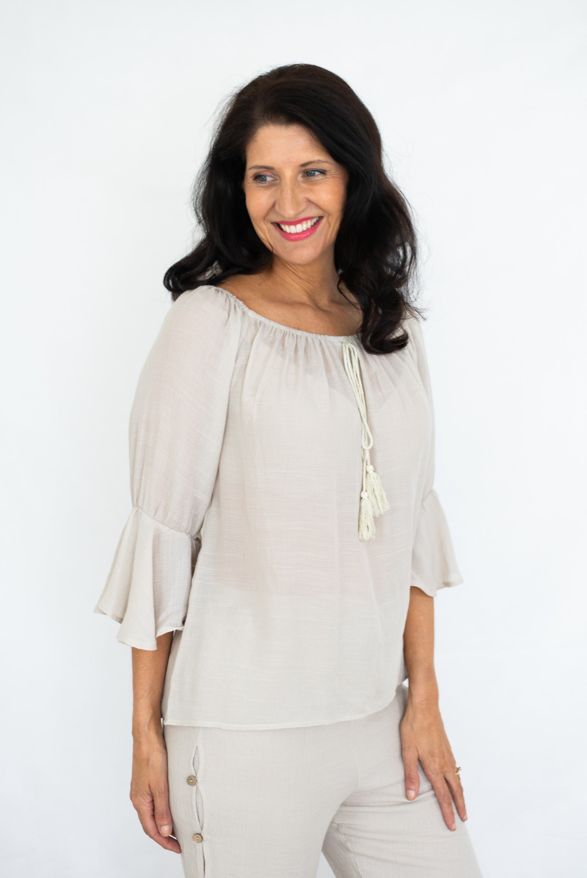 Woman in a beige Bell Top with 3/4 sleeves and tassel drawstring round neckline, and beige beach pants against a white background