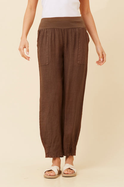 Woman wearing chocolate (or brown) Linen Italian Pants with an adjustable waistband, front pockets, and pleated hems, paired with a white top and white cross cross shoes. Cream studio background.