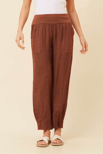Woman wearing rust coloured Linen Italian Pants with an adjustable waistband, front pockets, and pleated hems, paired with a white top and white criss-cross shoes. Cream studio background.