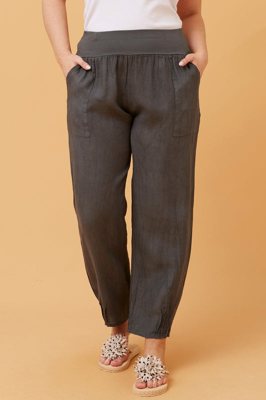 Woman wearing charcoal Linen Italian Pants with an adjustable waistband, front pockets, and pleated hems, paired with a white top and fluffy patterned shoes. Orange studio background.